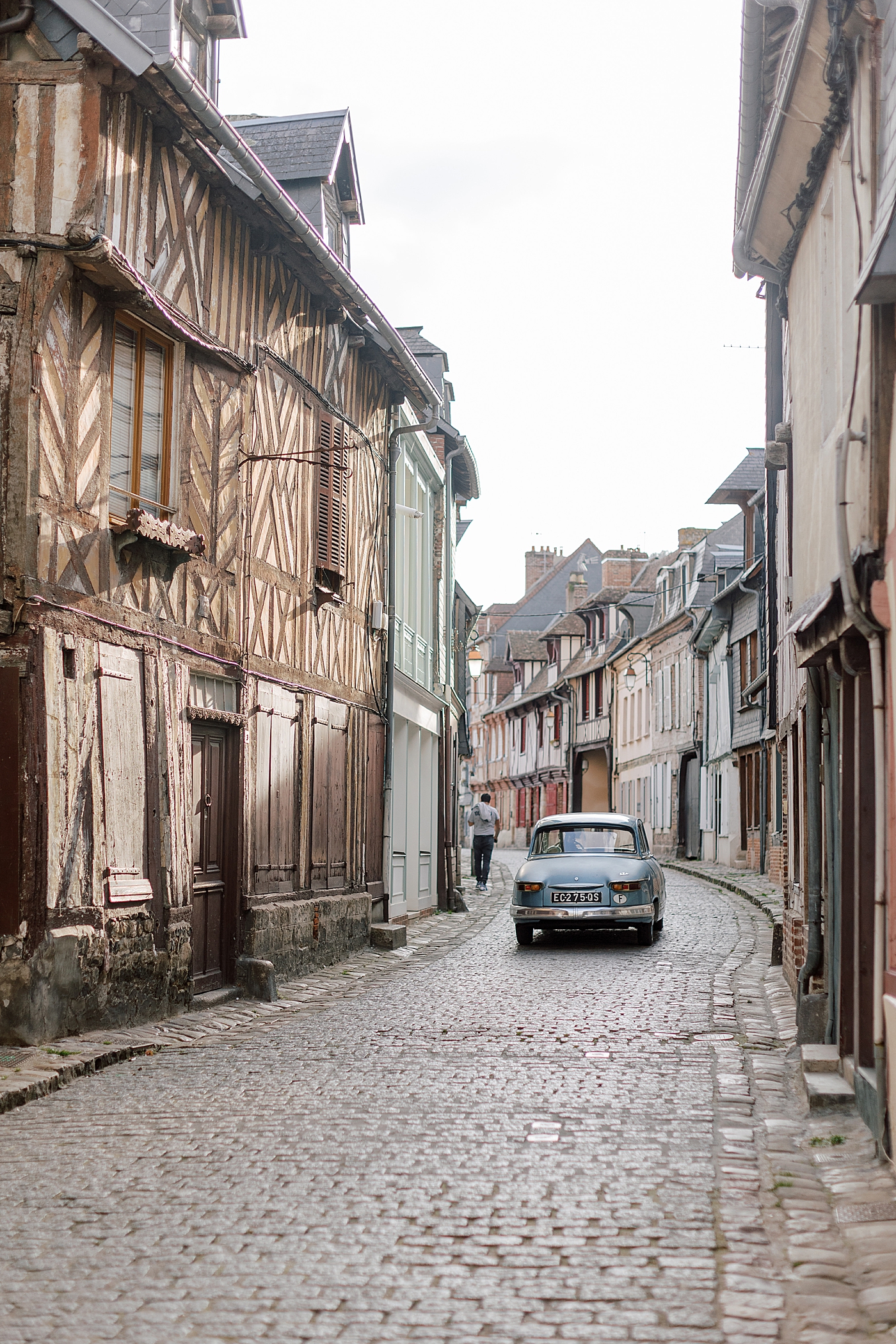 View of a French cobblestone street with homes, a car driving away, and a man walking during a trip with France Wedding Photographer Hope Helmuth Photography