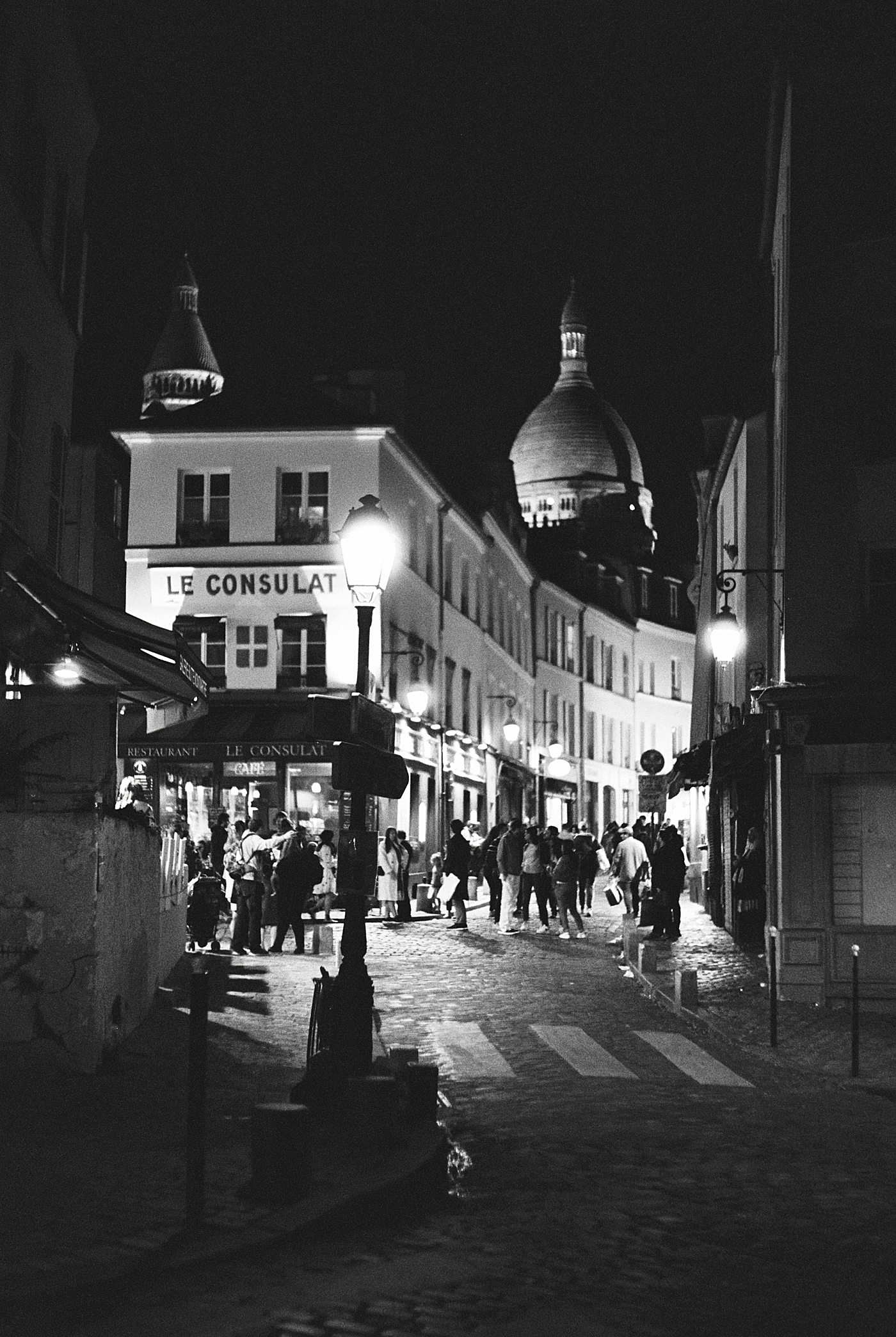 Black and white image at night of a cobblestone street full of people and street lamps | Image by Hope Helmuth Photography