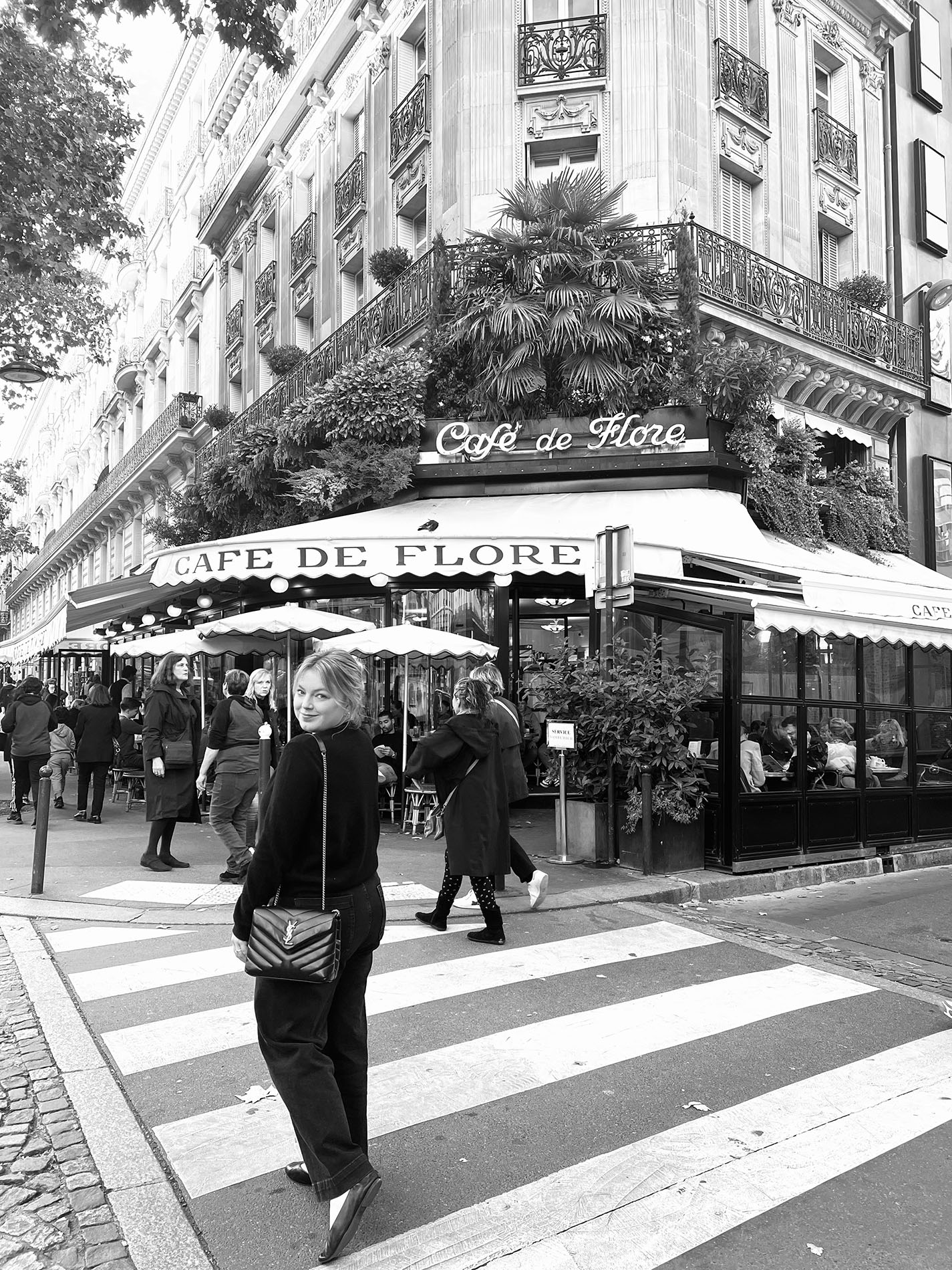 Black and white photo of a woman in an all black outfit and a loose bun walking across a crosswalk in France while looking back over her shoulder. 