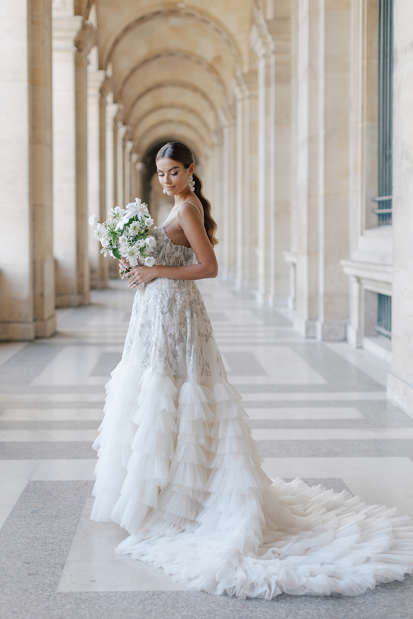 Bride standing in the outside hallway of the Louvre with a bouquet looking down over her shoulder | Image by Hope Helmuth Photography