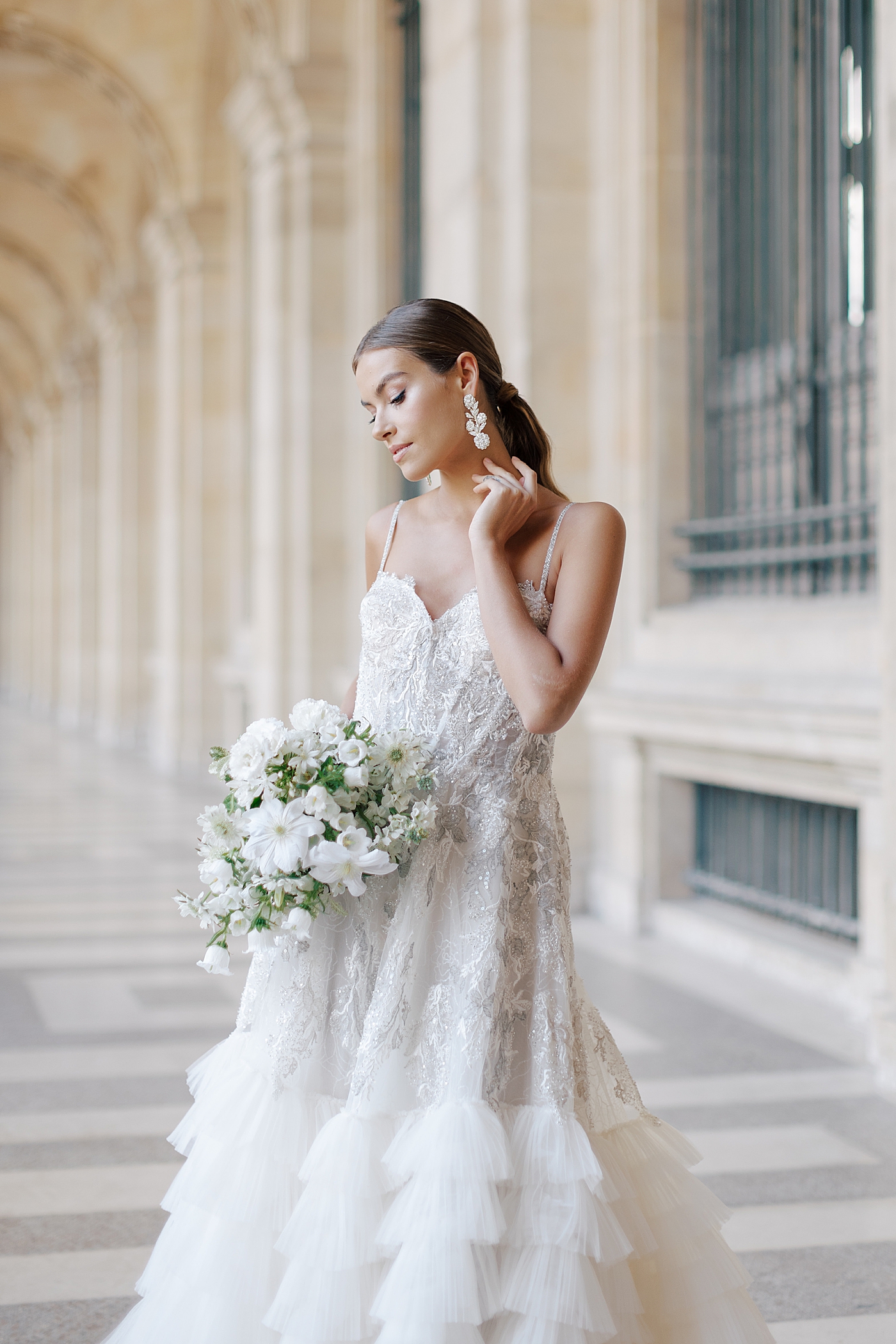 Bride standing in the outside hallway of the Louvre with a bouquet, holding her earring | Image by Hope Helmuth Photography