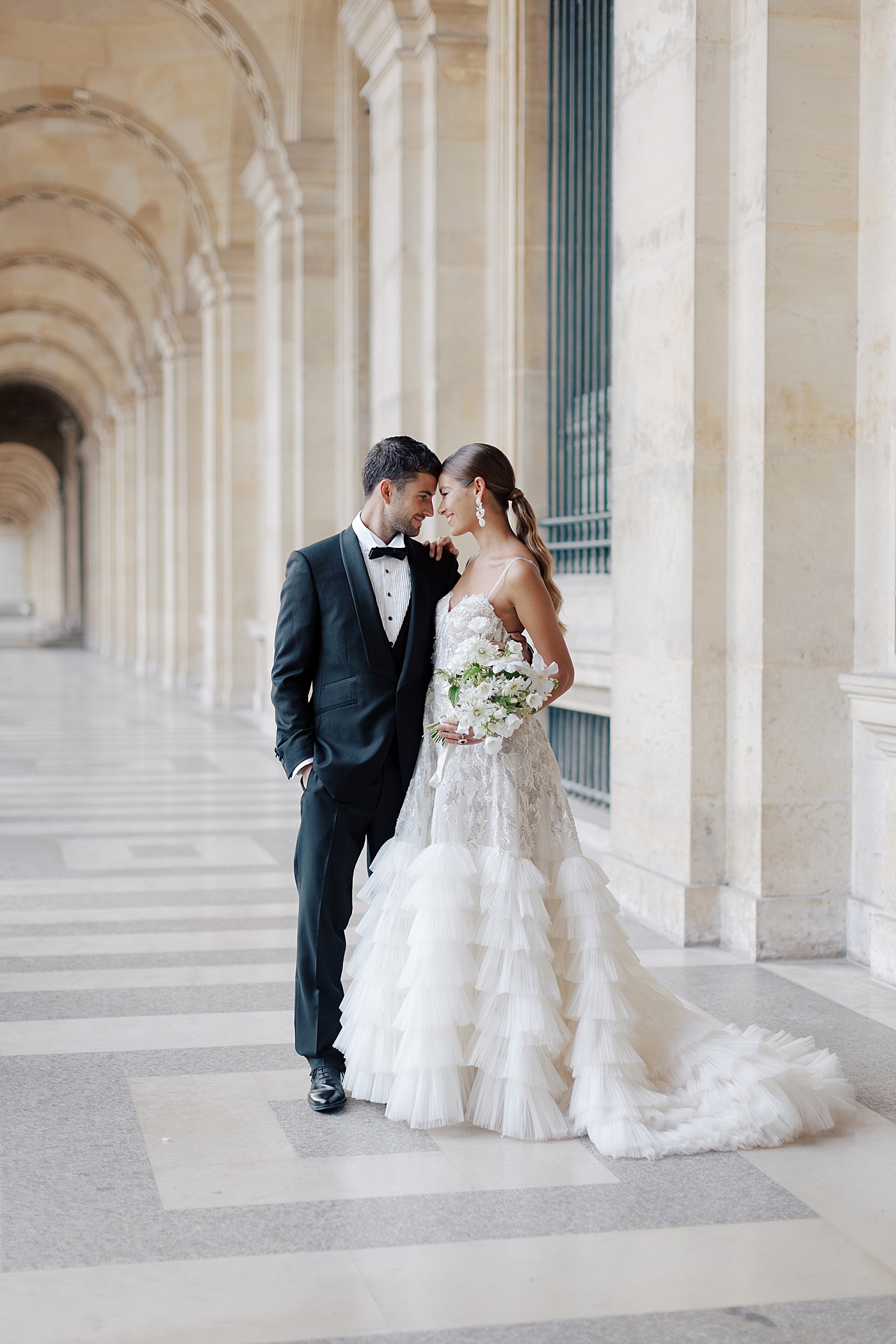 Bride and groom with their heads together standing in the outside hallway of the Louvre with a bouquet | Image by Hope Helmuth Photography