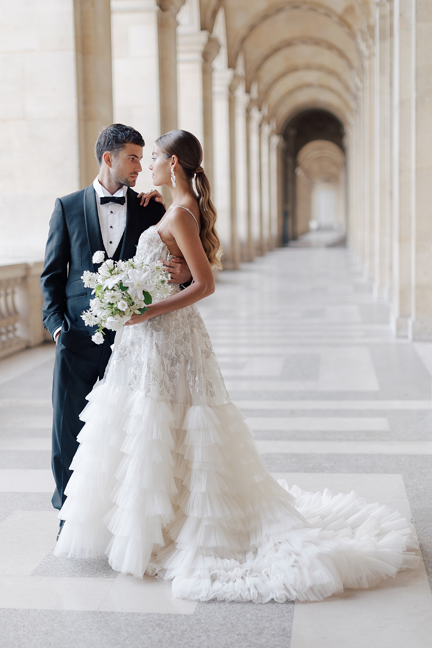 Bride and groom looking at each other while standing in the outside hallway of the Louvre with a bouquet | Image by Hope Helmuth Photography
