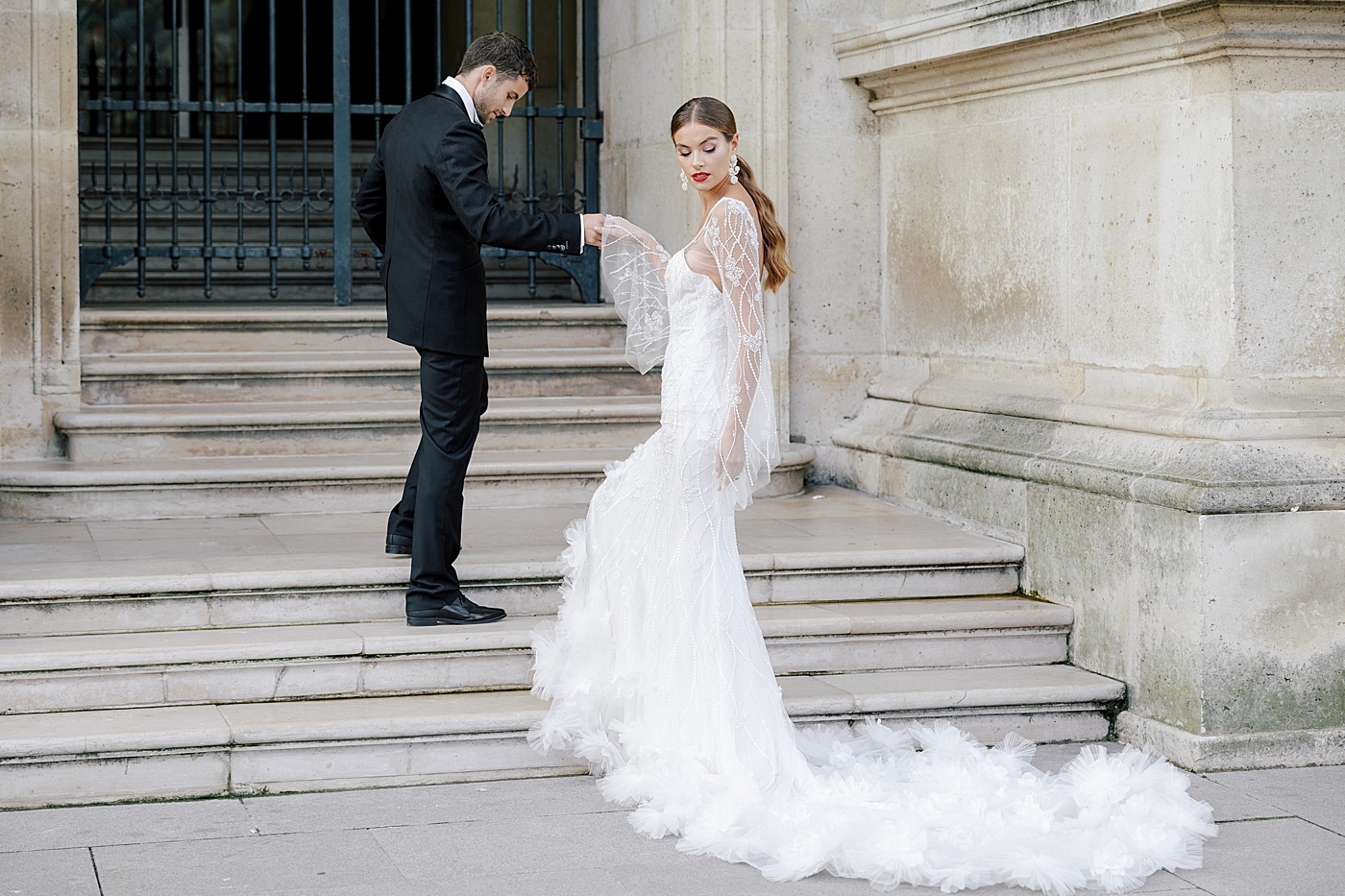 Groom leading his bride with a long, feathery train up concrete steps | Image by Hope Helmuth Photography