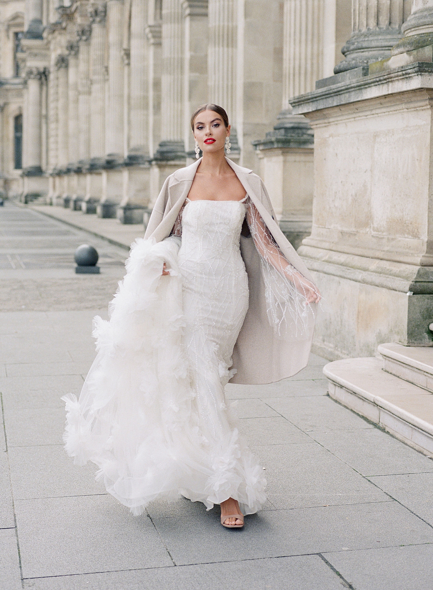 Bride walking towards the camera in a wedding dress with a train, beige coat, and bright red lipstick during Paris Elopement | Image by Hope Helmuth Photography